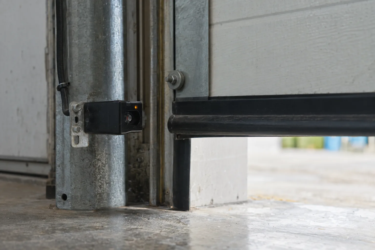 Close-up of a photo-eye safety sensor mounted on the side track of a commercial overhead door at a warehouse bay, with a safety reversing edge visible along the bottom of the door, lit by ambient daylight from outside.