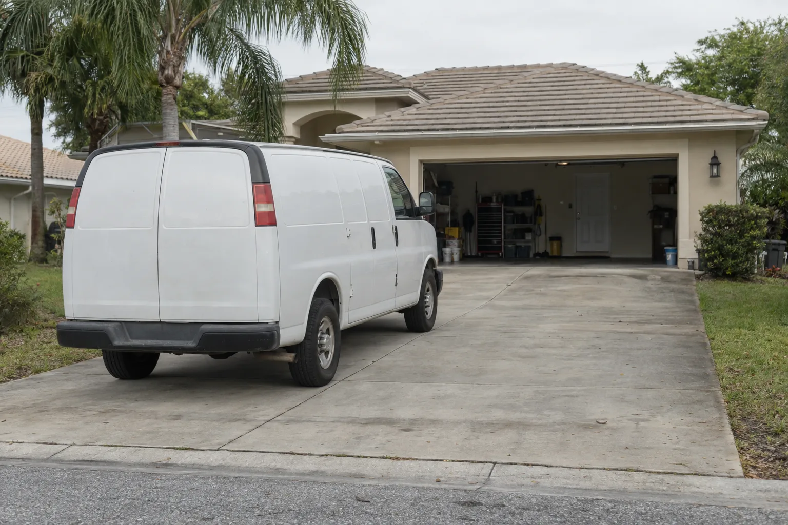 A white service van parked in a residential driveway in front of a stucco Florida home with an open two-car garage, palm trees visible on either side in bright morning sunlight