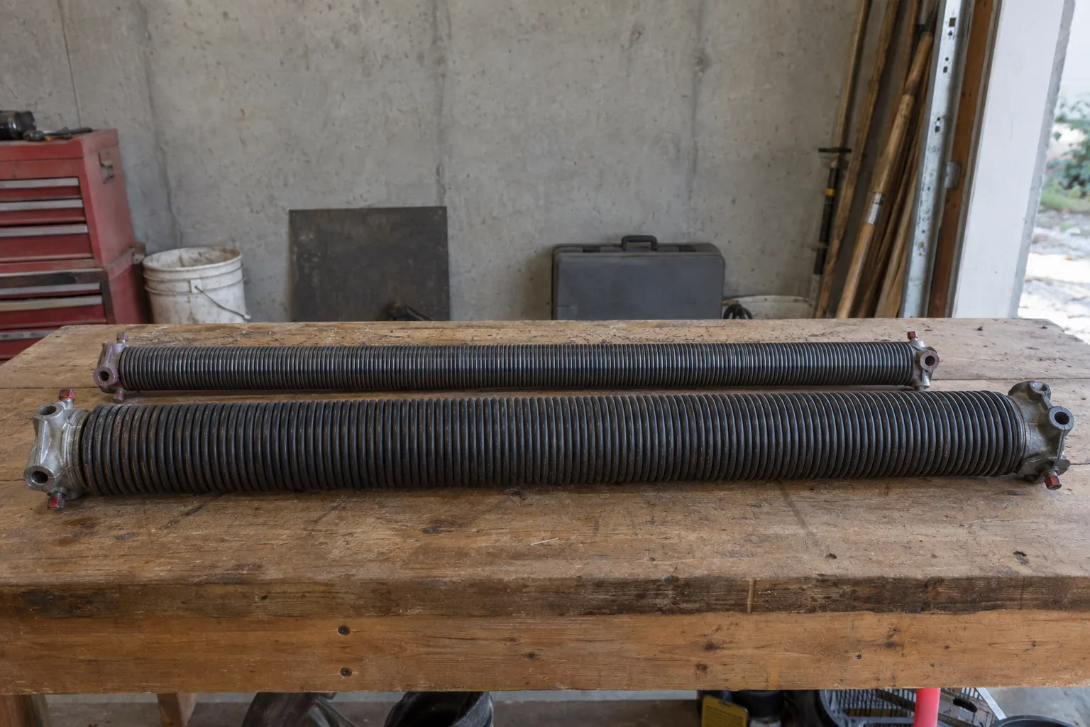 Two torsion springs of different wire gauges placed side by side on a wooden workbench under fluorescent workshop lighting, showing a clear size difference between the two coiled springs