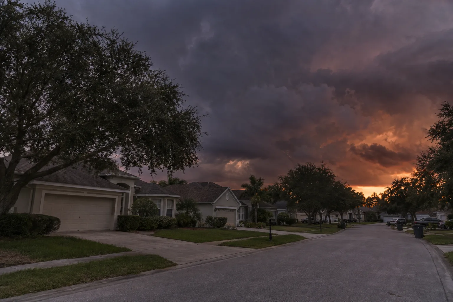 Dark storm clouds build at dusk over a Central Florida suburban street lined with stucco homes with two-car garages and live oak trees, an orange glow from the setting sun visible on the horizon