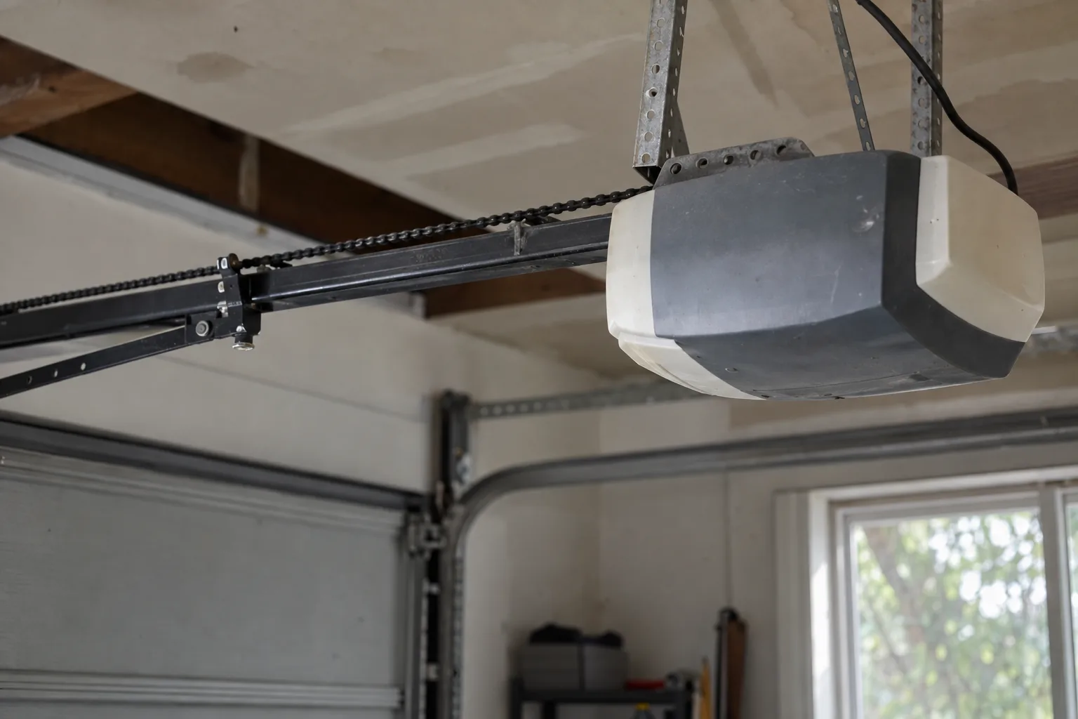 Residential garage door opener motor unit mounted on a ceiling rail, viewed from below at a low angle inside a suburban Florida garage, with a belt drive and trolley arm visible under natural daylight