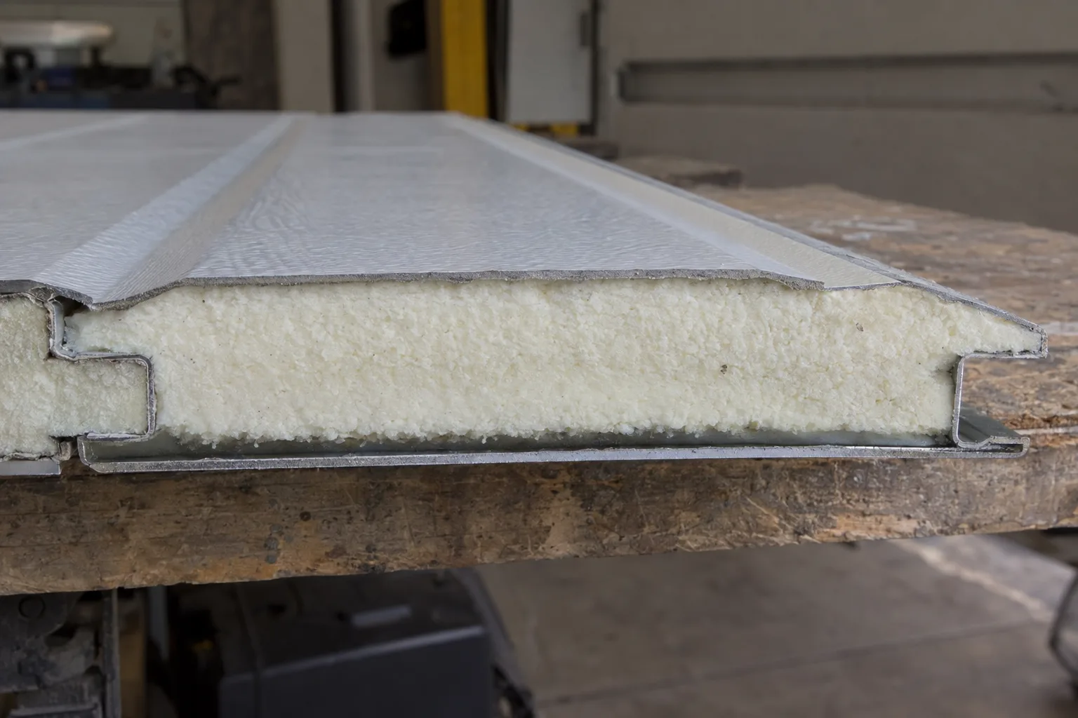 Cross-section of a garage door panel resting on a workbench, showing a thick foam insulation core pressed between two steel skins, illuminated by warm workshop lighting