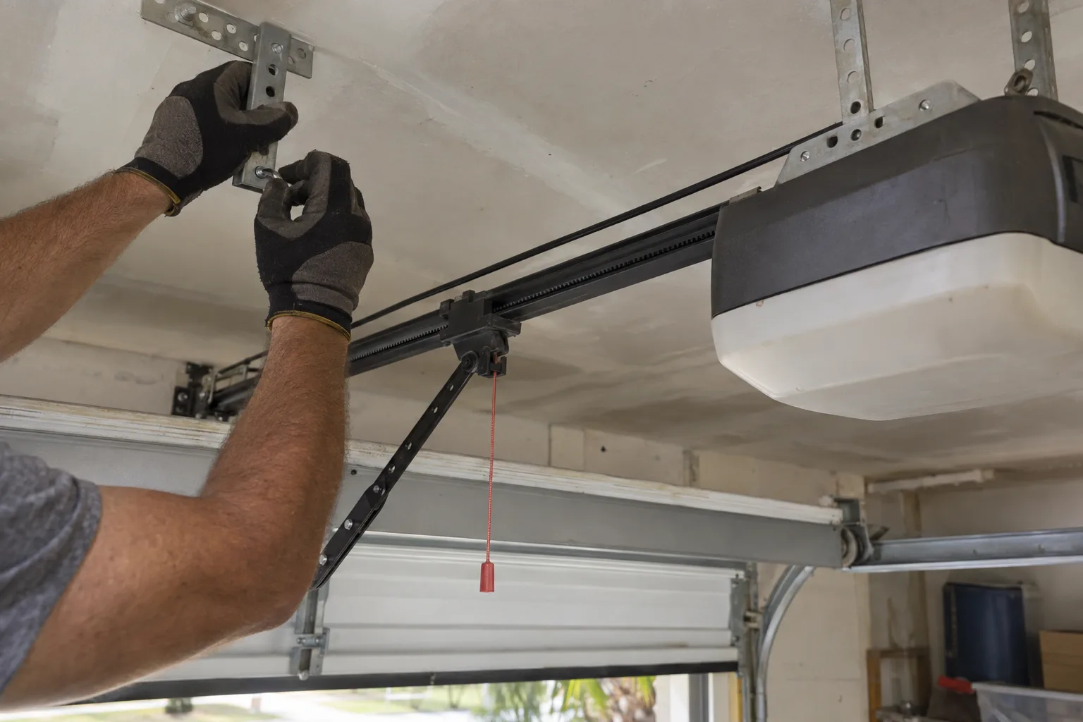 A technician's gloved hands securing a belt drive garage door opener rail to a white garage ceiling, with the garage door visible below and natural daylight filling the space
