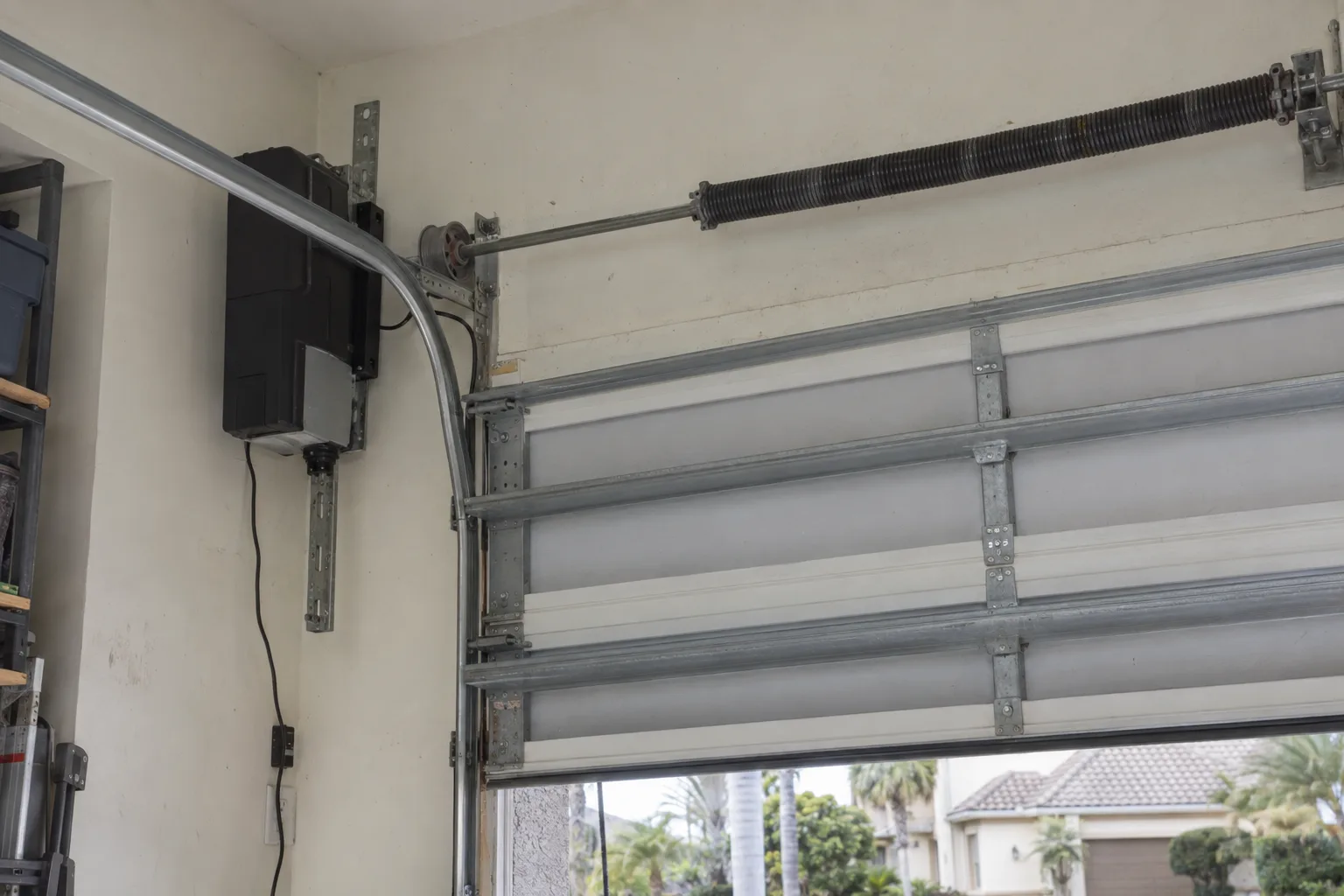 A wall-mounted jackshaft garage door opener installed on a side wall next to the horizontal torsion shaft and spring assembly above a closed sectional garage door inside a bright residential garage