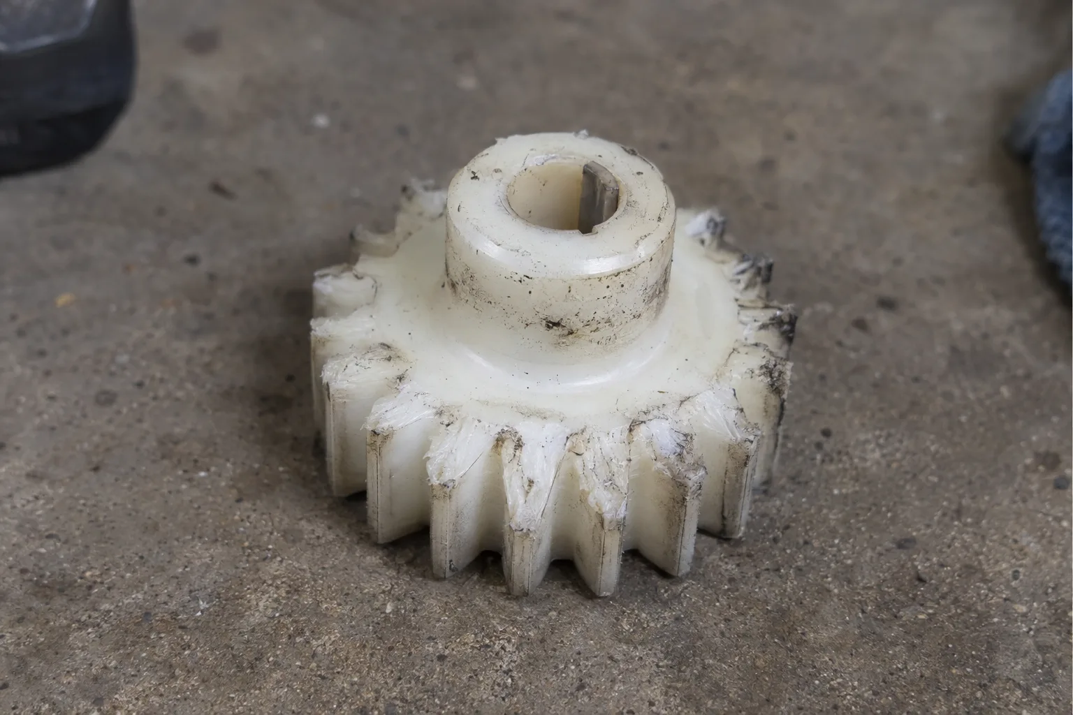 A worn white plastic drive gear with visibly stripped teeth removed from a garage door opener, resting on a concrete surface in close-up detail