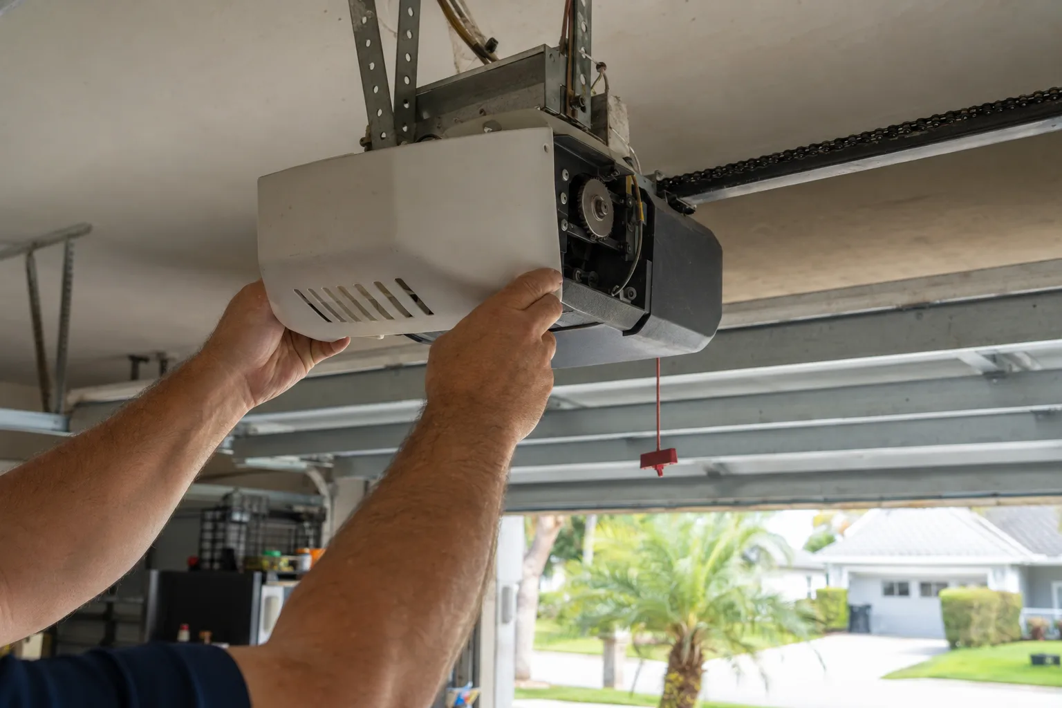 A technician's hands removing the cover panel of a ceiling-mounted garage door opener inside a residential garage, with natural daylight coming through the open garage door behind