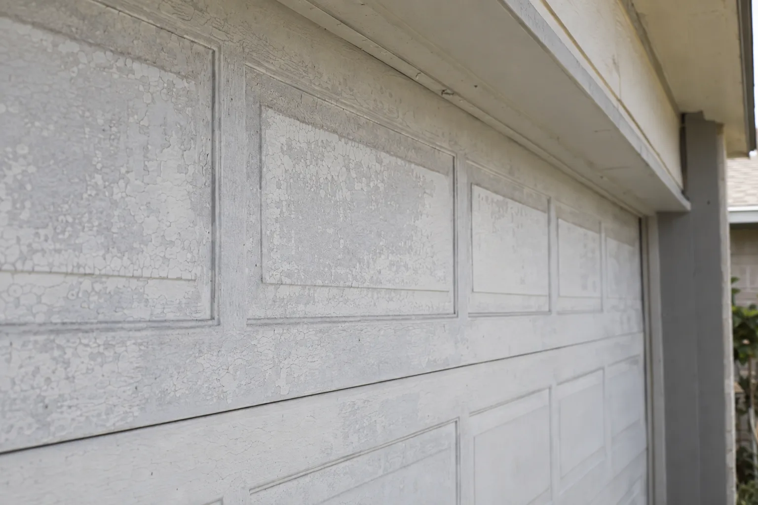 Upper panels of a garage door showing chalky, faded, and lightly cracked paint degradation under harsh afternoon sunlight, with surface texture clearly revealed by strong directional light