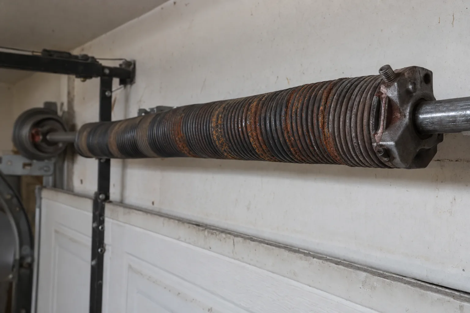 Close-up of a torsion spring above a residential garage door with visible rust and corrosion forming along the steel coils, lit by natural daylight inside a Florida garage.