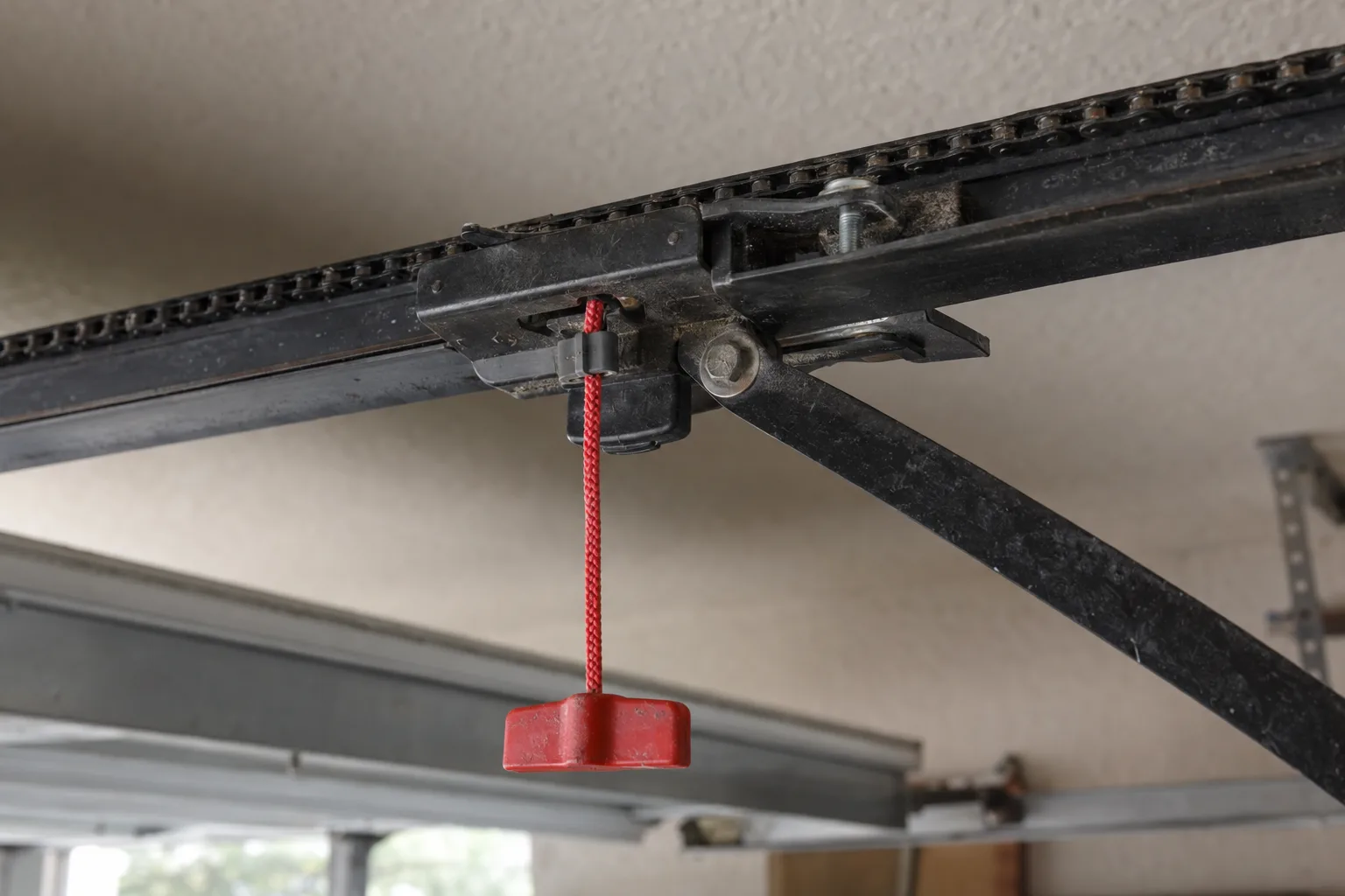 Close-up of a garage door opener trolley carriage with a red manual release cord hanging down, dimly lit residential garage interior with soft natural light from above