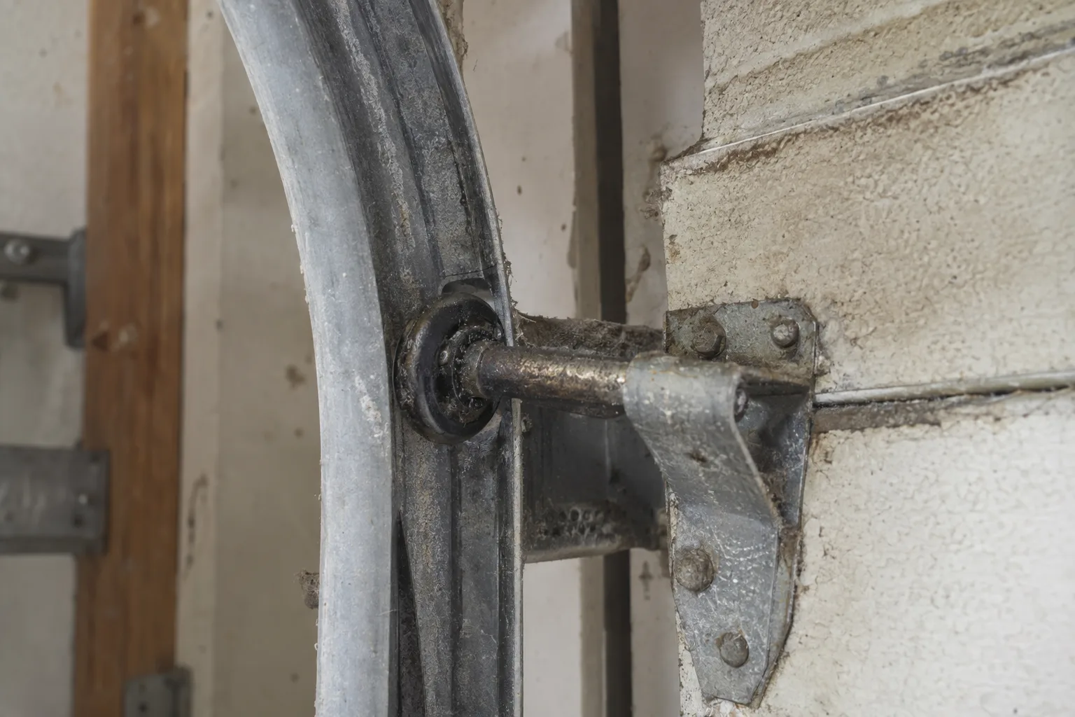 A steel garage door roller and track section heavily coated in dust and dried lubricant buildup, photographed up close inside a residential garage with strong sidelighting that highlights the grime and wear.