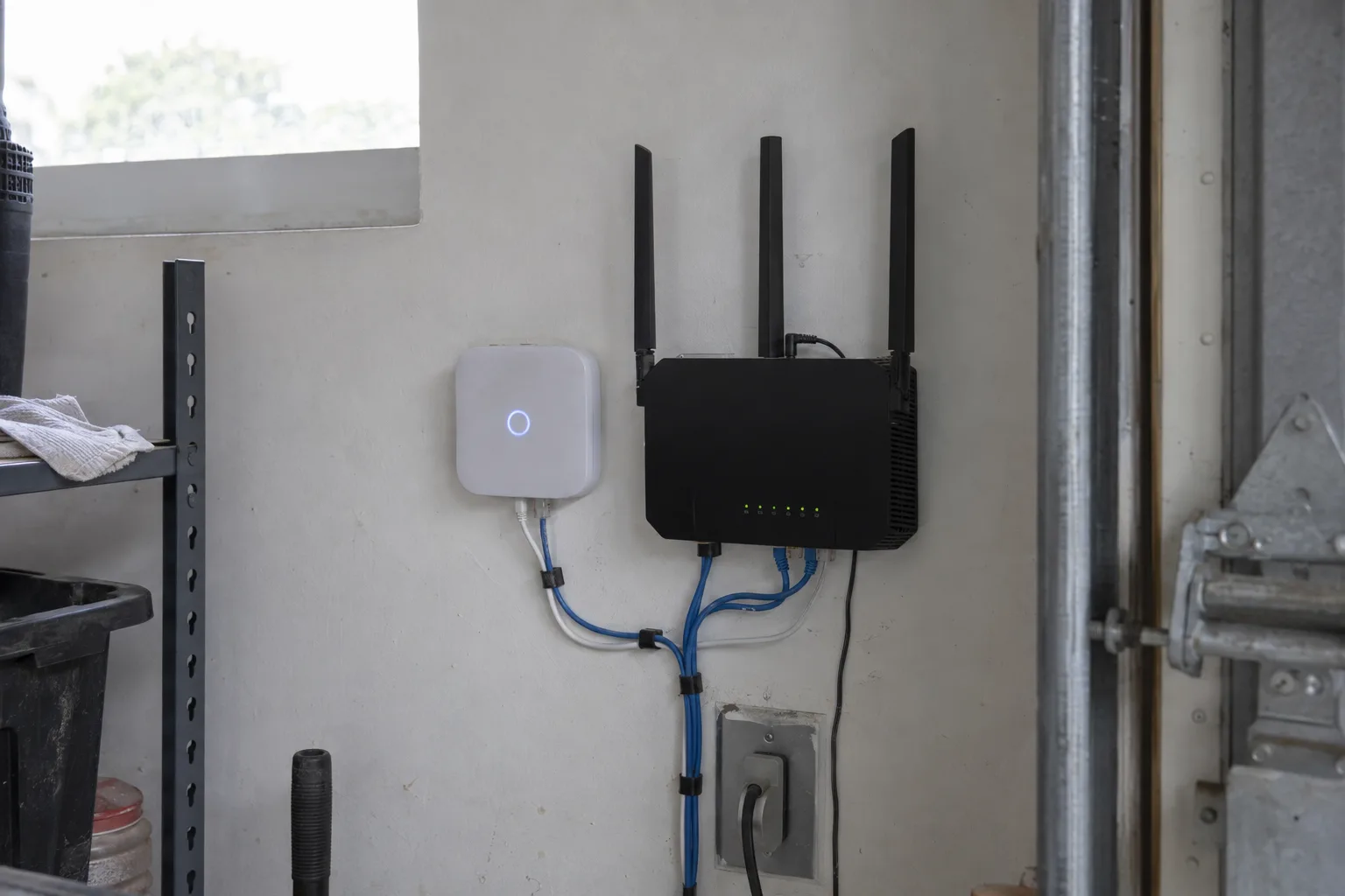 A smart home hub and Wi-Fi router mounted side by side on a white garage wall, with indicator lights on and cables neatly routed, lit by natural daylight from a nearby window