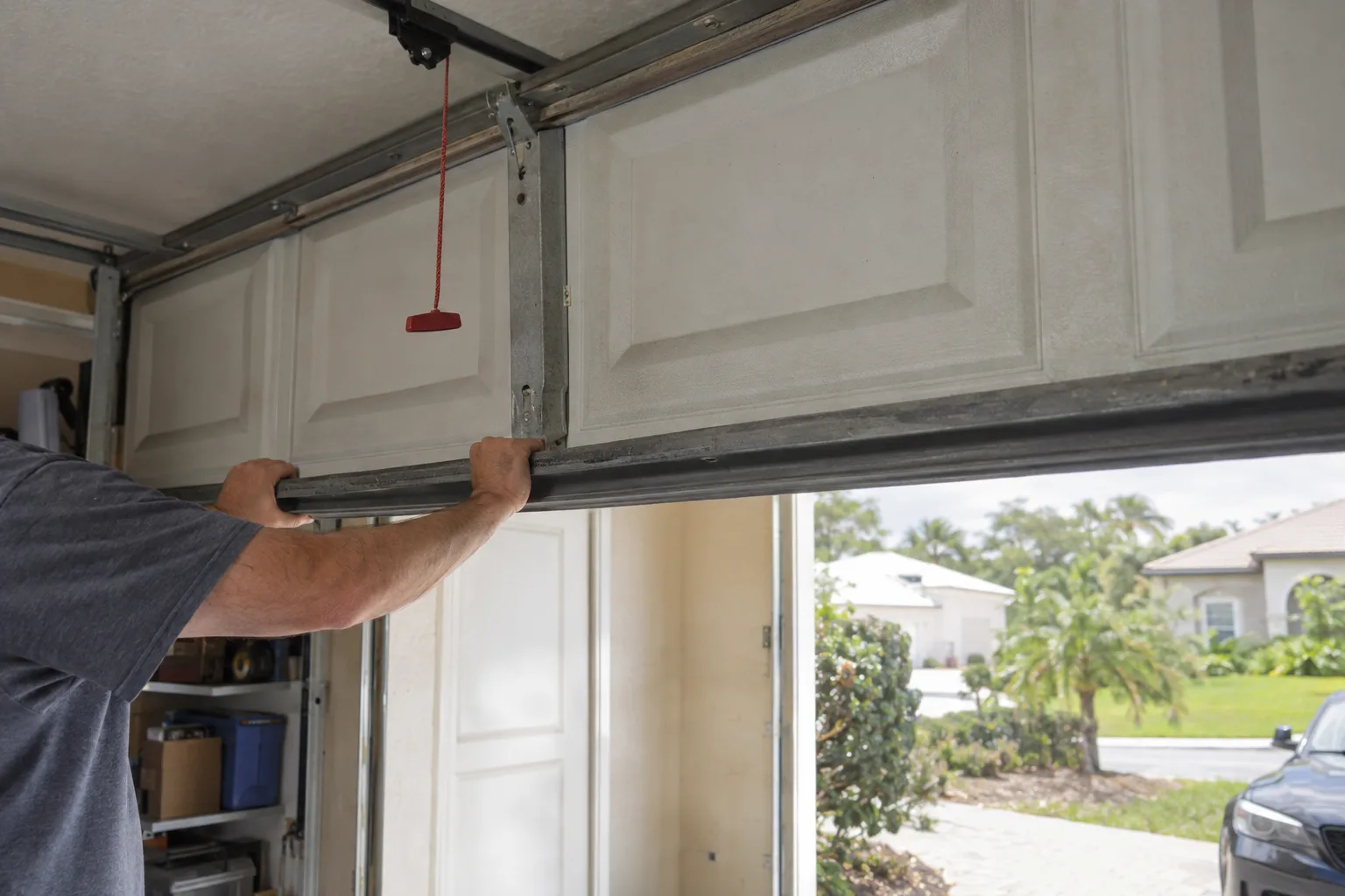 A technician's hands gripping the bottom rail of a sectional garage door, pulling it manually along its tracks inside a sunlit residential garage with the emergency release cord hanging loose from the opener trolley.