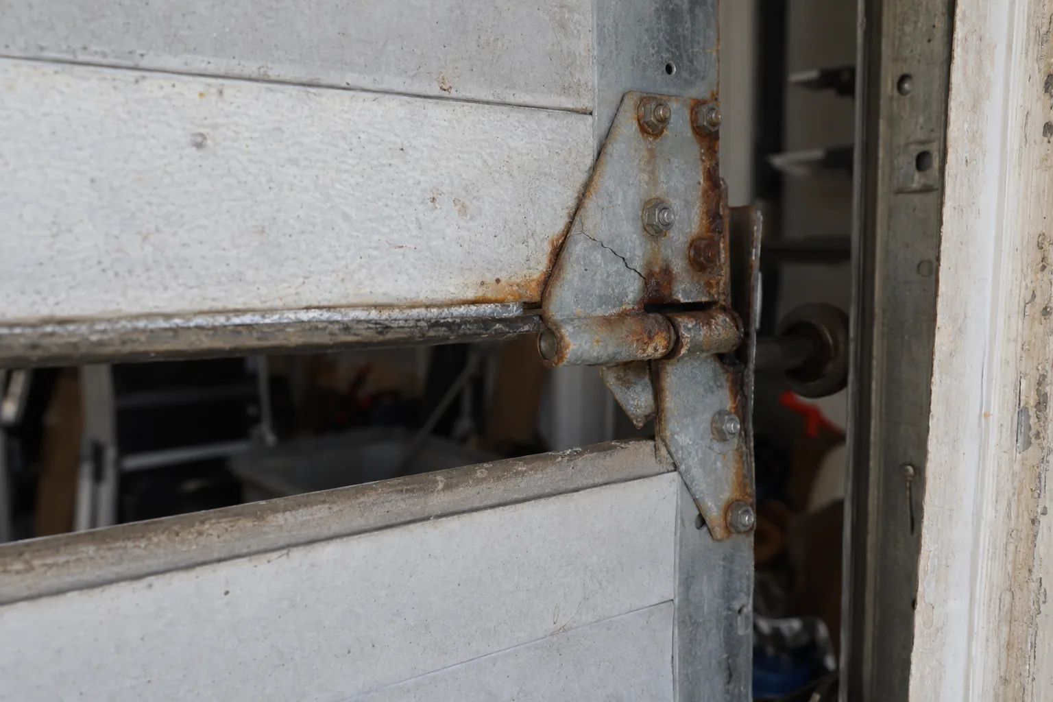 Close-up of a cracked, rust-pitted steel garage door hinge at a panel joint, with the door slightly raised and the garage interior dimly visible behind it