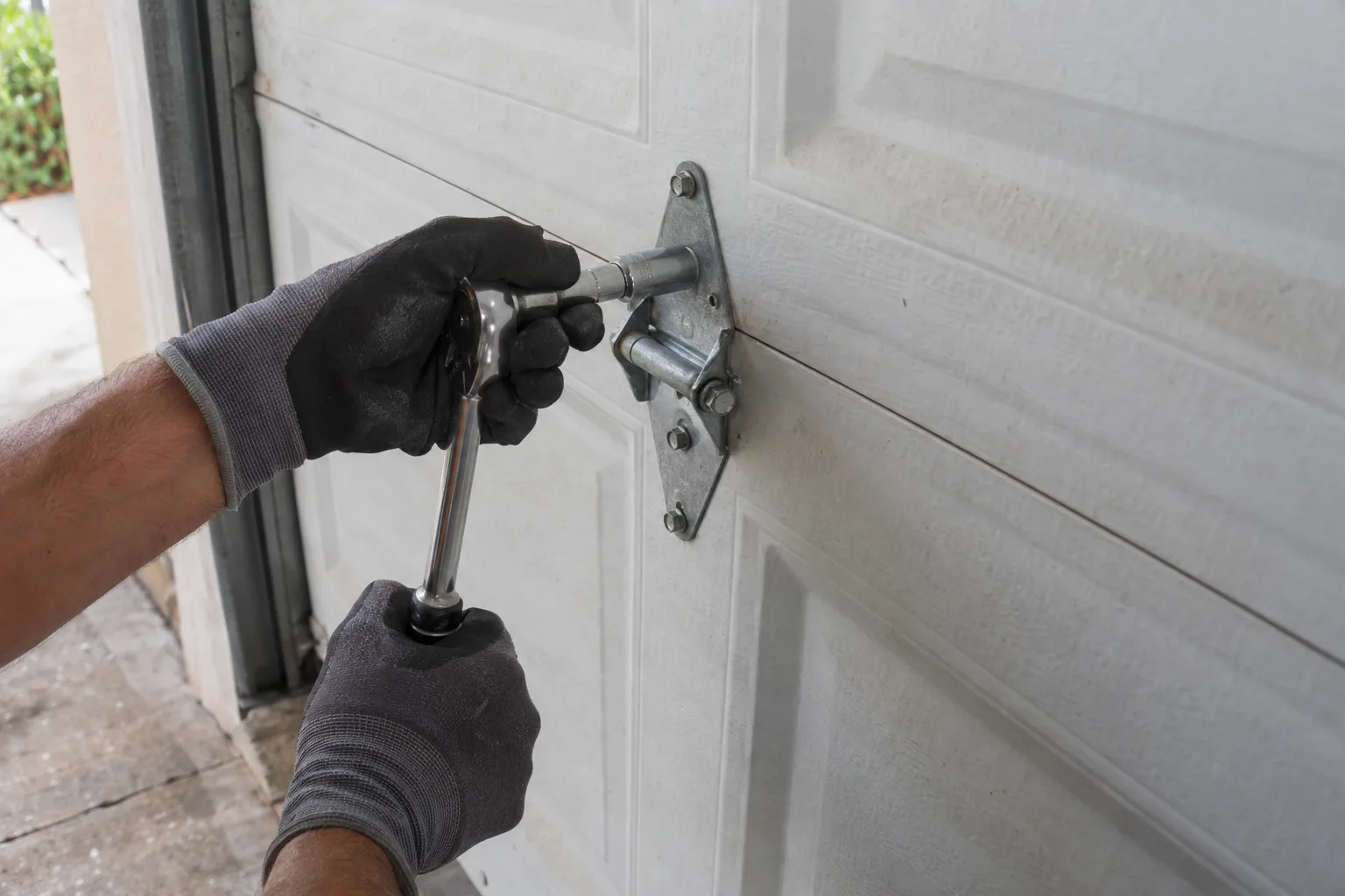 Gloved hands using a socket wrench to fasten a steel hinge between two panels of a white sectional garage door in a sunlit Florida driveway