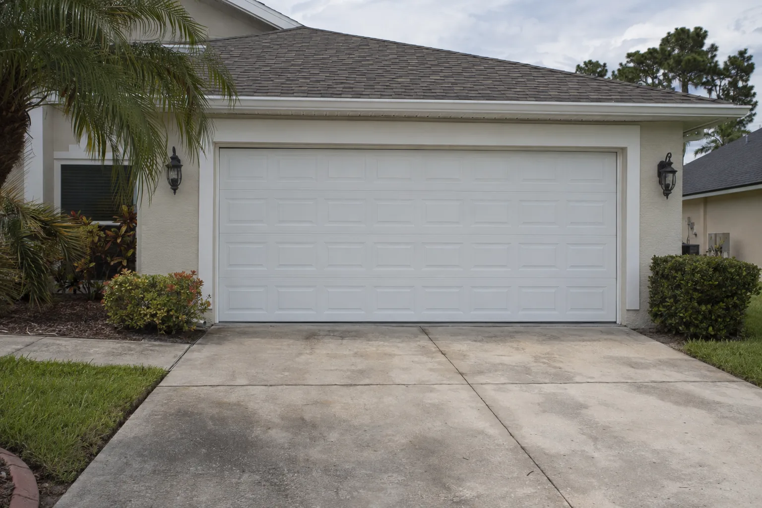 A two-car garage with a newly installed white insulated sectional door on a pale stucco suburban Florida home, surrounded by green grass and tropical plantings under midday sun.