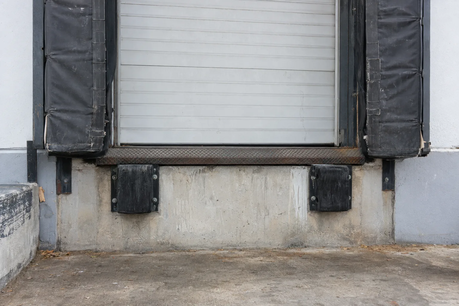 A warehouse loading dock bay face showing a foam dock seal with canvas covering flanking the opening and two black rubber bumper pads bolted to the concrete apron below