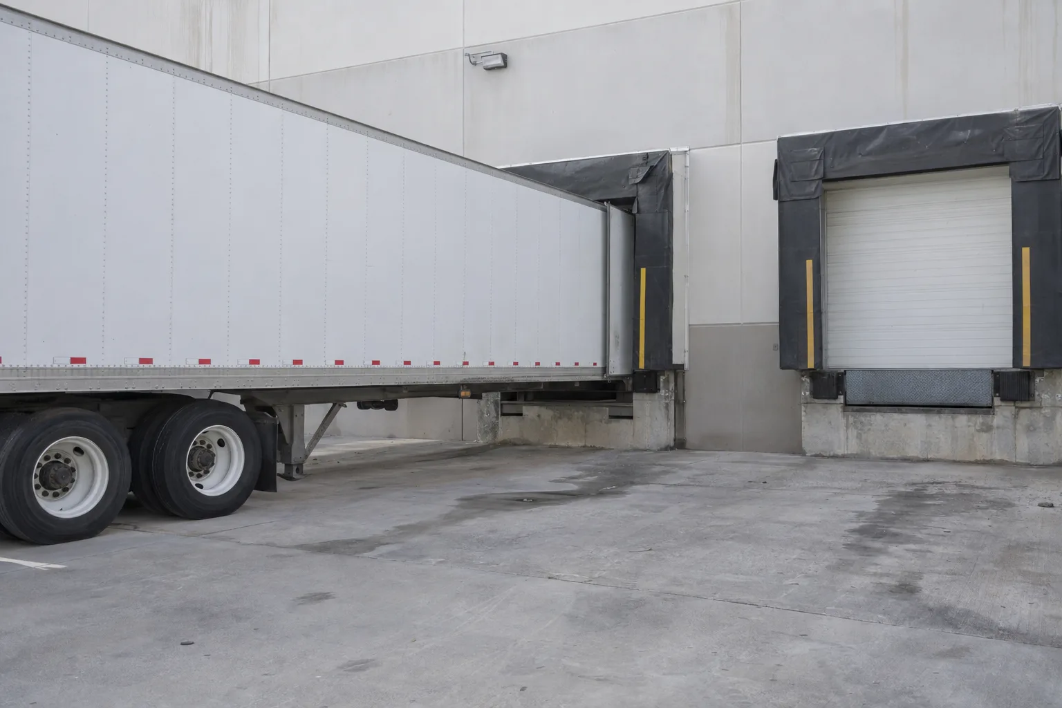 A white semi-trailer backed into a warehouse loading dock with the steel dock leveler plate deployed and bridging the gap between the dock and the trailer floor in late-afternoon sunlight