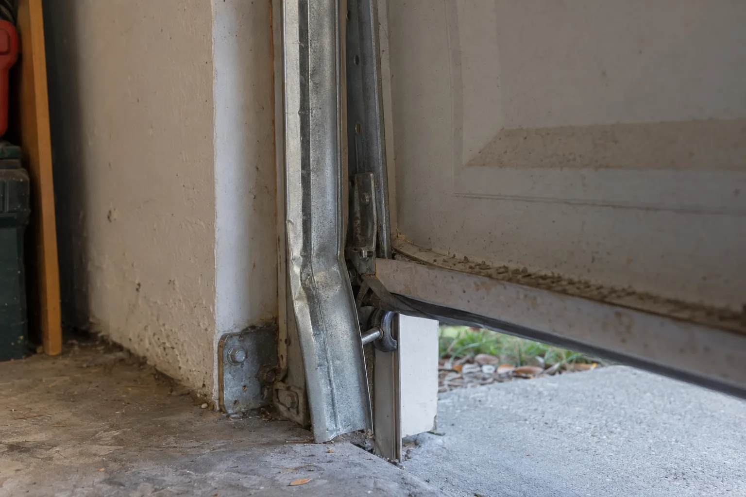 A bent and kinked garage door track section viewed up close inside a garage, with a misaligned door panel partially separated from the rail and a displaced roller visible near the bracket