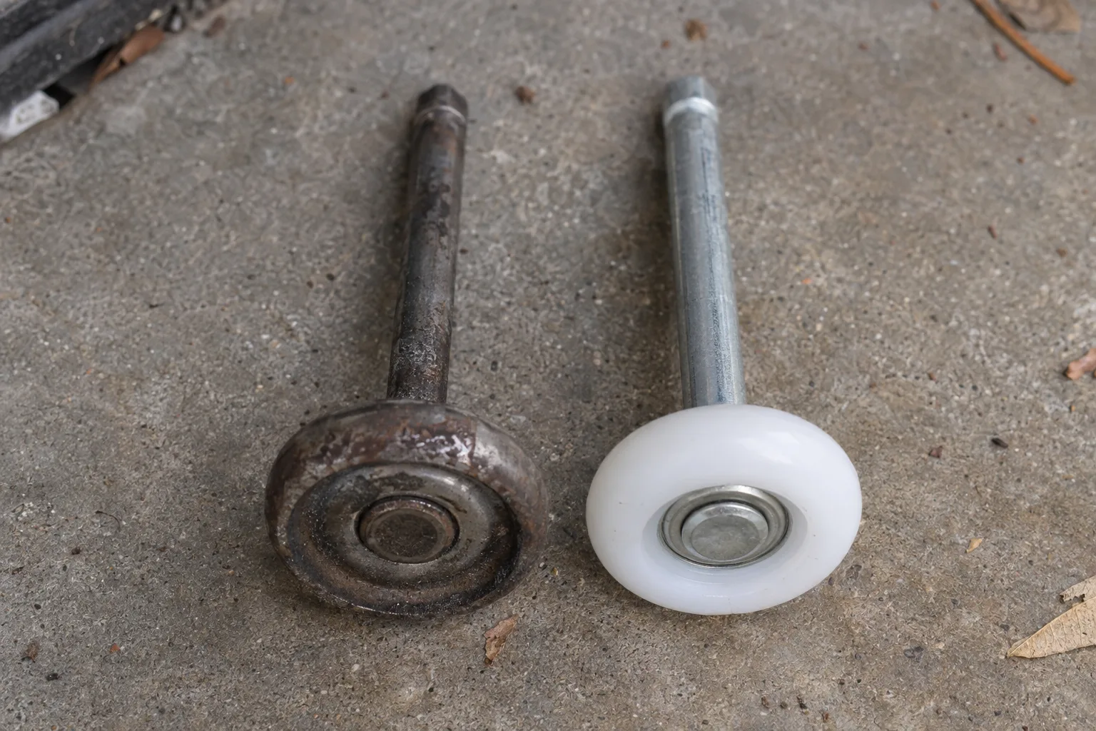 A worn steel garage door roller with a discolored rusty stem placed beside a new white nylon roller on a concrete garage floor