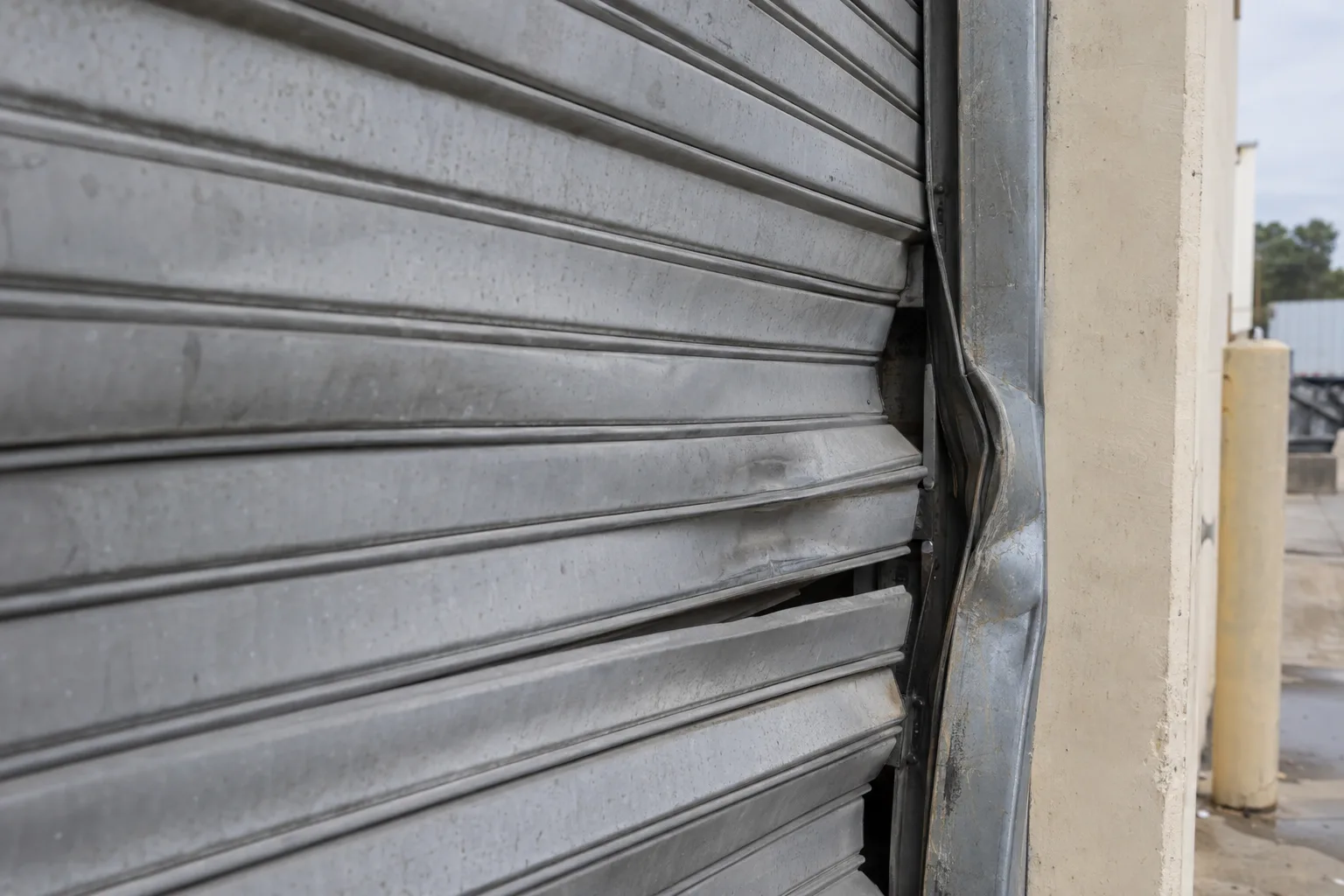 Bent and dented steel slats on a commercial rolling door with a visibly misaligned and warped side guide rail, photographed in flat natural daylight against a concrete warehouse wall.