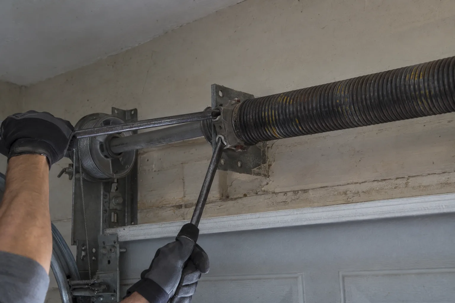 A gloved hand shining a flashlight onto a garage door torsion spring and track bracket in a dimly lit residential garage