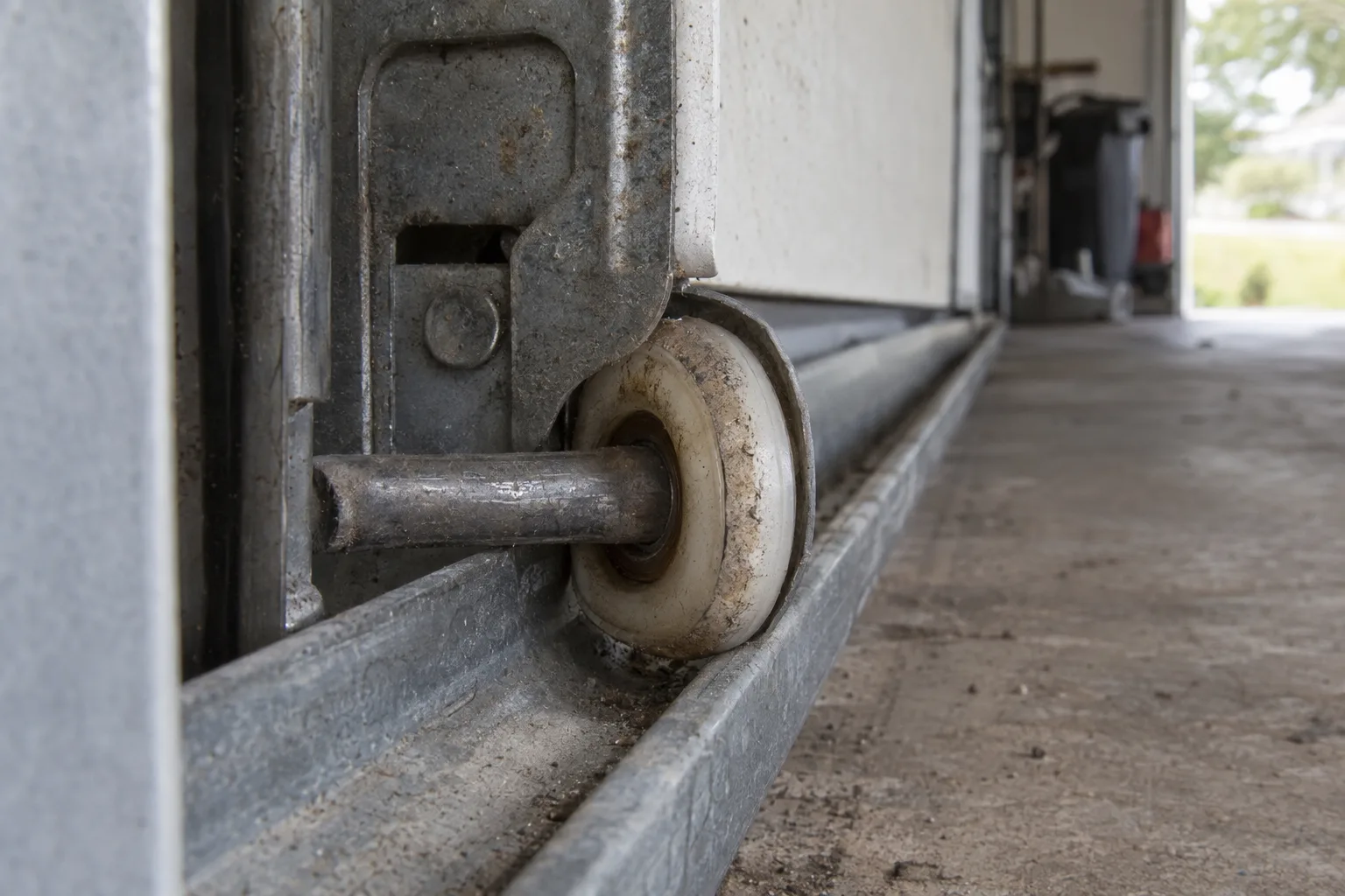 Worn garage door roller with a flattened stem inside a galvanised steel track, photographed at track level inside a residential garage with soft natural light