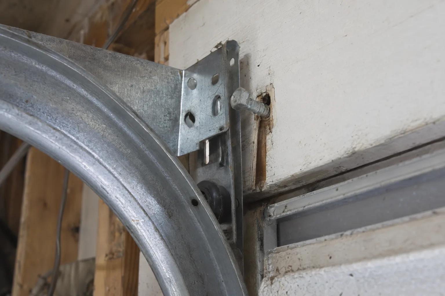 Close-up of a garage door track mounting bracket with a partially backed-out lag bolt, showing the bracket pulled away from a wooden header board inside a garage