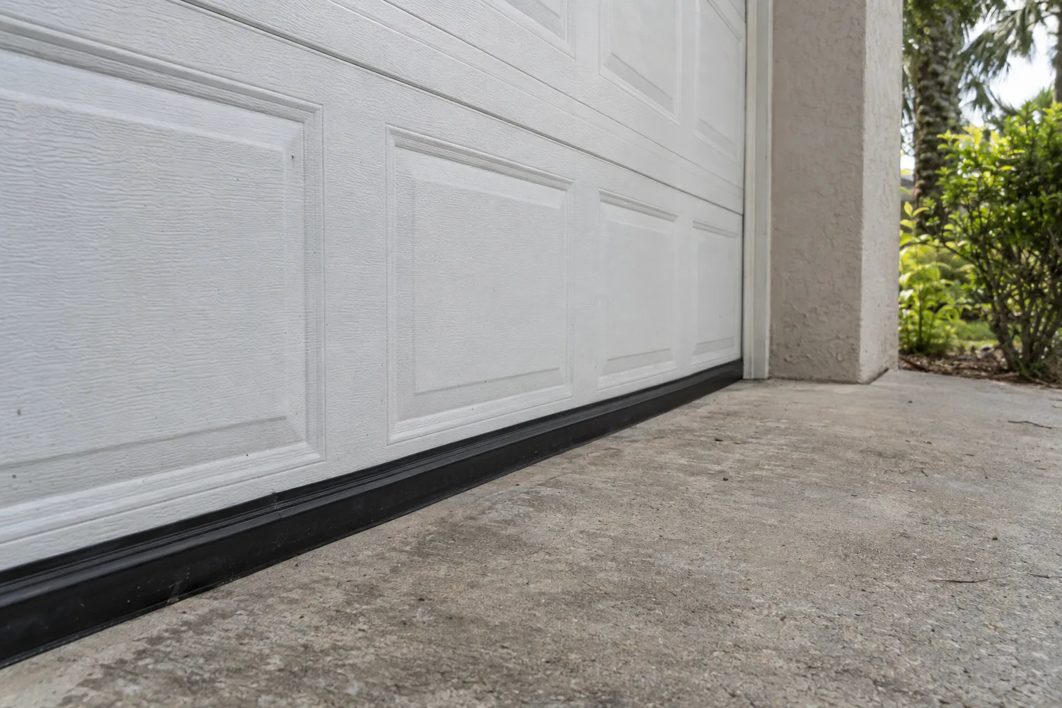 A new black rubber bottom seal fitted snugly along the base of a white garage door on a sunlit concrete driveway, with soft green subtropical vegetation visible in the blurred background