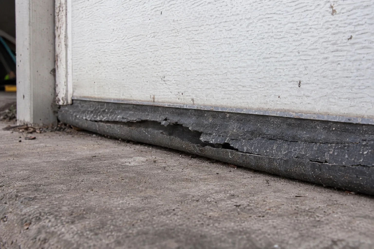 A hardened and cracked rubber garage door bottom seal in poor condition resting against a concrete slab, with visible splits and gaps caused by sun and heat deterioration